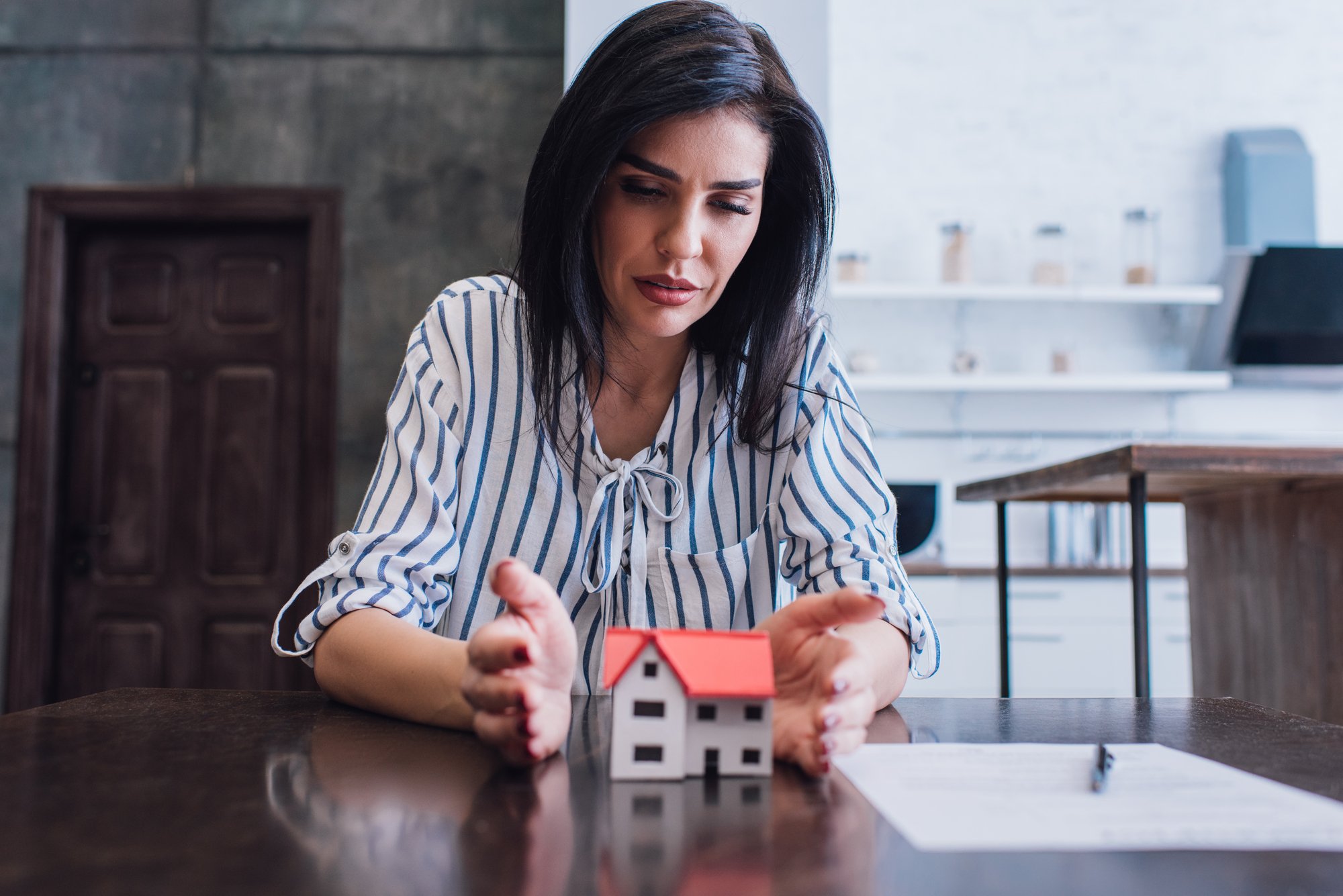 Concentrated woman putting hands near house model with paper and pen on table in room