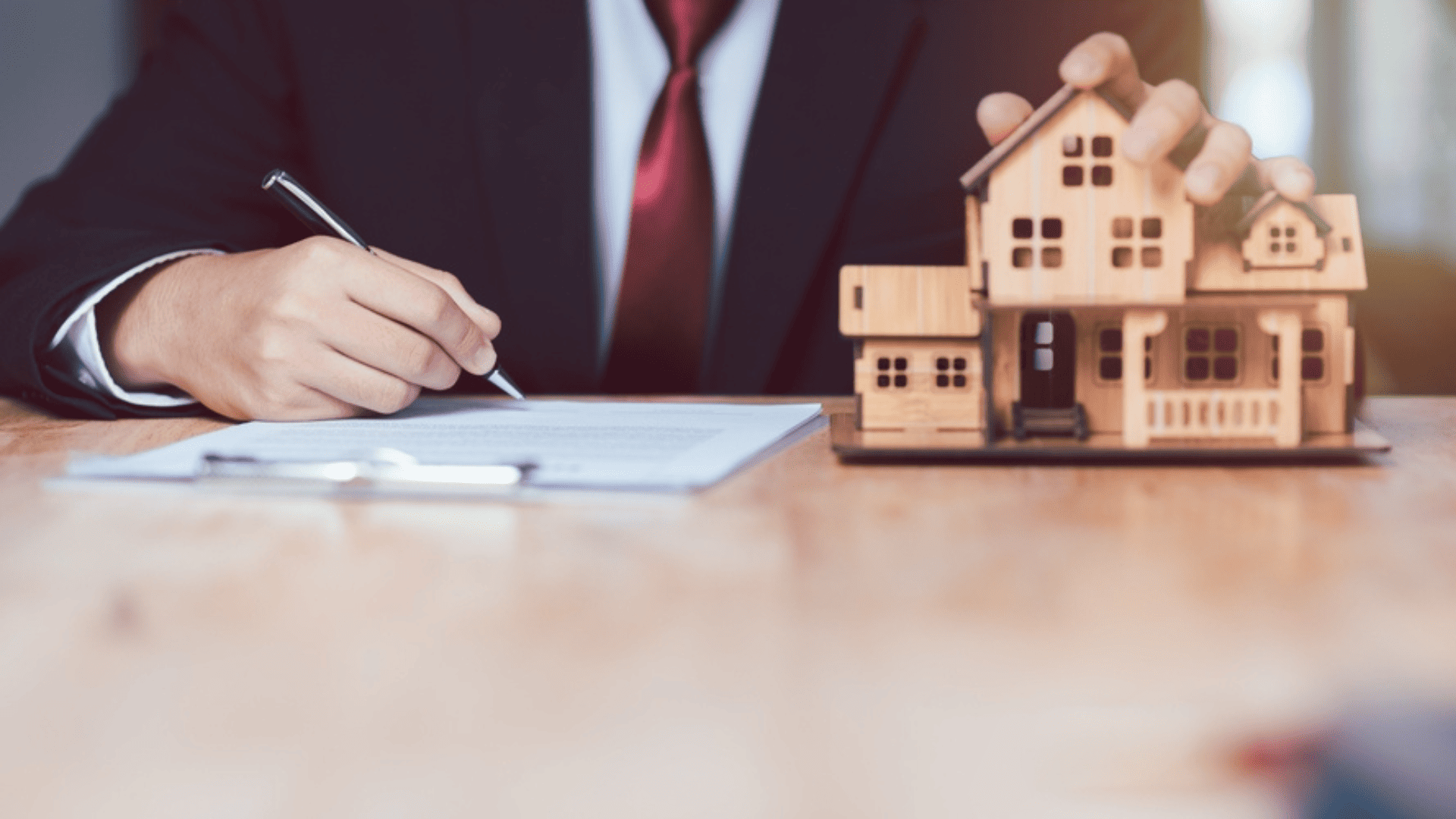 Businessman signing document with one hand while other hand rests on wooden house signaling protection of the rental asset.