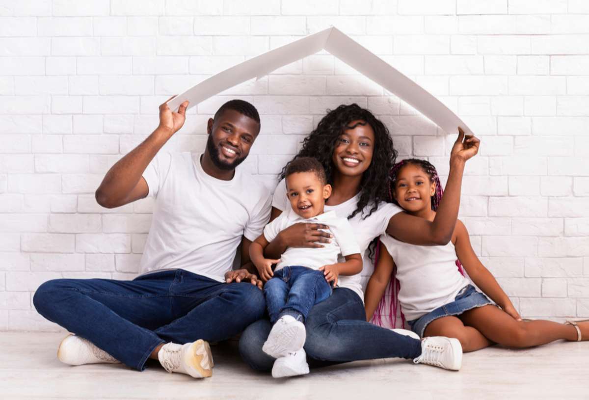 Young African American Parents With Two Children Sitting Under Cartboard Roof Dreaming Of New Home