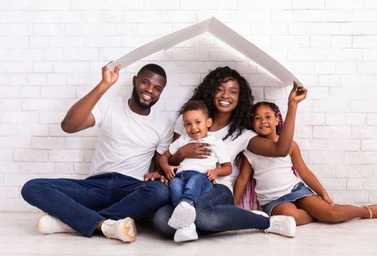 Young African American Parents With Two Children Sitting Under Cartboard Roof Dreaming Of New Home