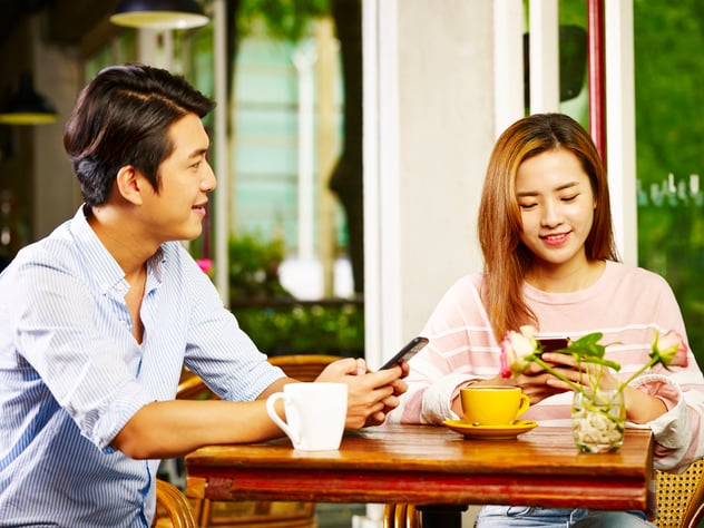 Young asian man and woman sitting at table chatting talking playing with cellphone in coffee shop or tea house