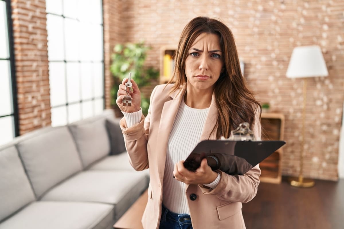 Young brunette real state agent woman holding keys of a home, frowning upset because of problem