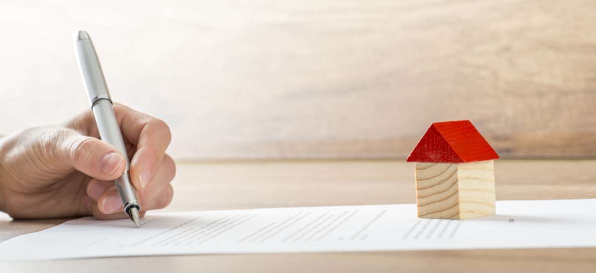 Closeup of new homeowner signing a contract of house sale or mortgage papers with a wooden toy house on the document